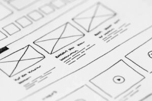Web Design Sheffield: close-up of a hand-drawn website wireframe on paper showing a grid of rectangular placeholders marked with “X,” lines representing text blocks, and a circular play-button icon.